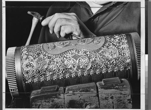 Black and white photograph of a metal cylinder covered in floral patterns being worked by a pair of hands with a small hammer