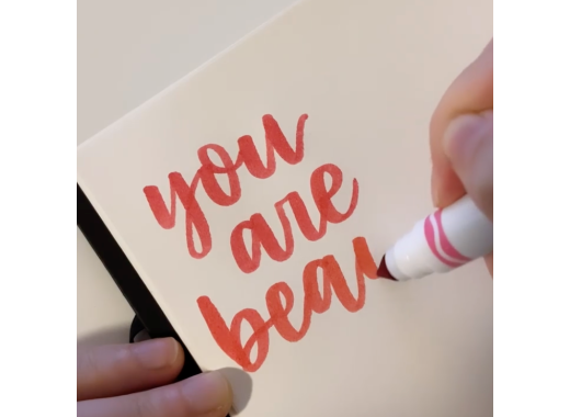 Photo of calligraphic words in pink marker reading "you are bea". Words are on white paper and a white hand is holding a pink Crayola marker.