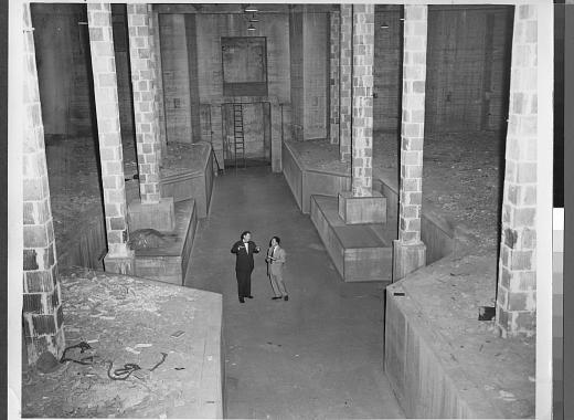 Black-and-white photograph of two men standing in the center of an unfinished basement with columns along each side