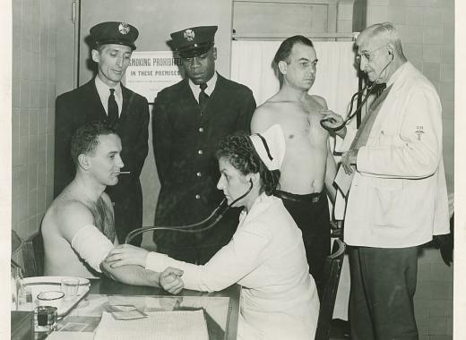 Black and white image depicting a nurse in the foreground with a stethoscope on the arm of a man. In the background, two men stand with FDNY seals on their hats. On the righthand side, a standing doctor holds a stethoscope to the heart of a man