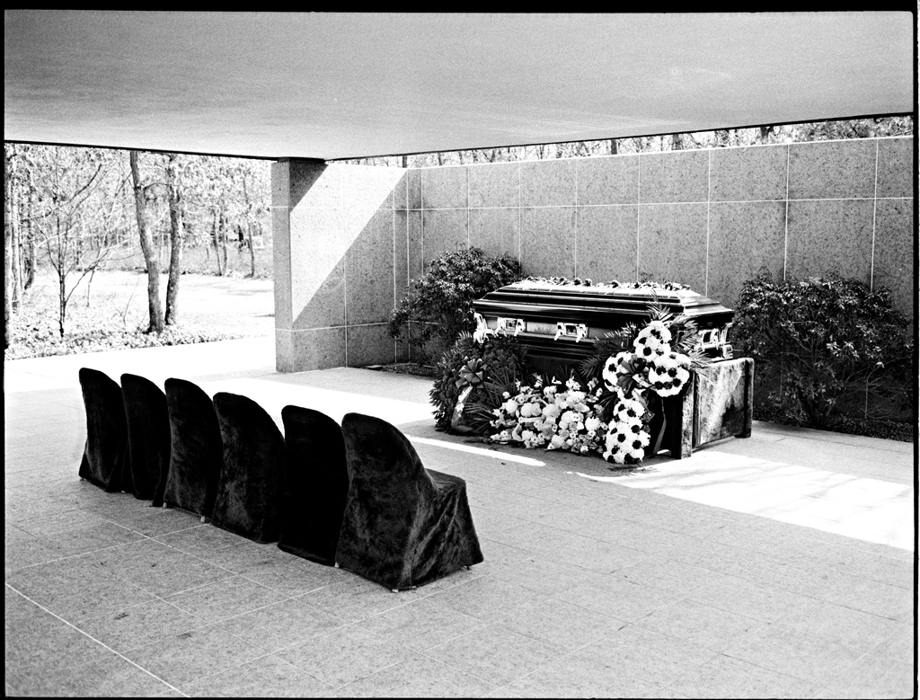 Photo of a coffin covered in flowers and 4 chairs draped in black at a cemetery.