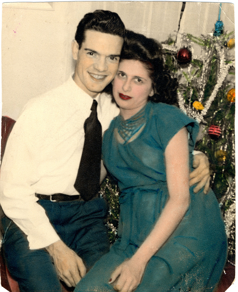 Photo of man and woman with dark hair smiling and embraced in front of Christmas tree. The woman is wearing a green dress, the man a white shirt and black tie.