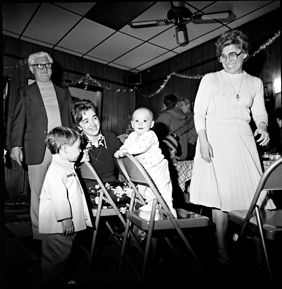 Smiling family sitting and standing in a decorated room. One smiling baby is standing on a chair while a toddler looks on