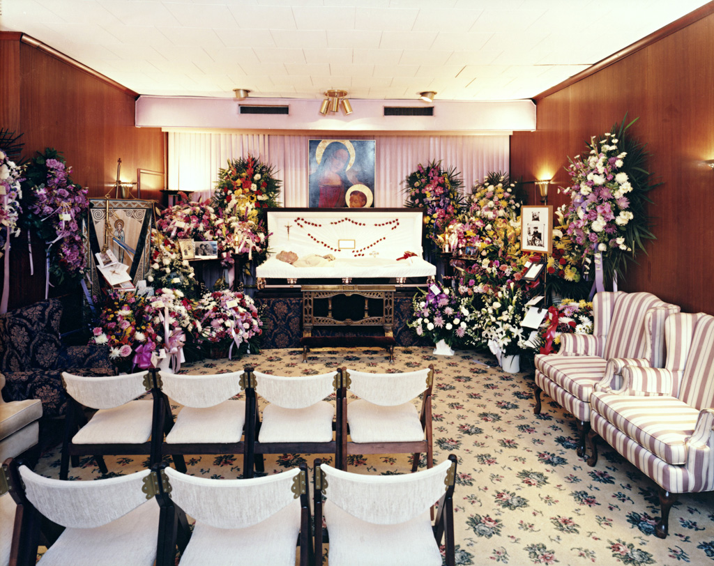 Photo of an open casket in a funeral parlor surrounded by large wreaths.