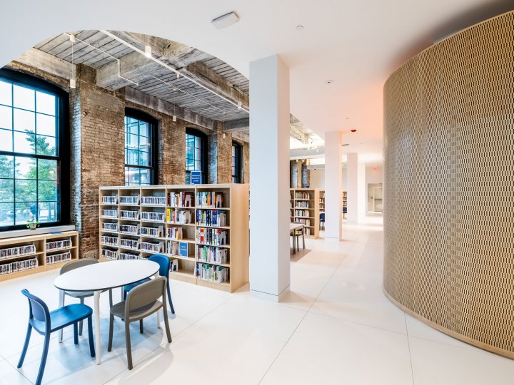 An interior photograph of Adams Street Library. The right side of the photo is a curved wall and on the left are bookshelves next to a pillar. In the lower left hand corner are a table and chairs.