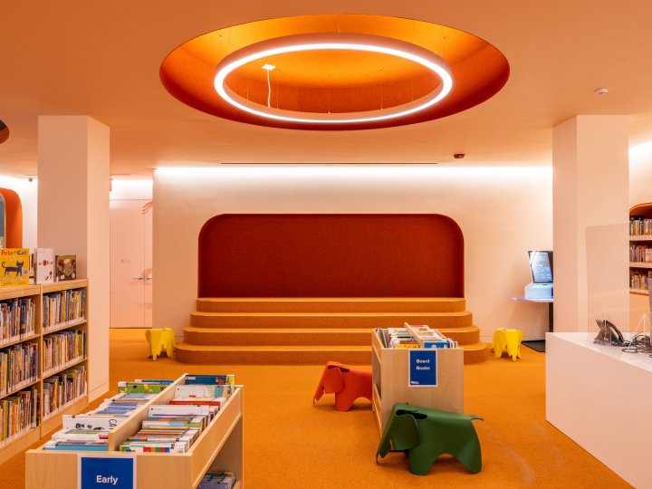 Adams Street Library's children's section: The room is a light orange color with an orange carpet. There are low tables and chairs that look like elephants in the middle of the floor. At the back of the room are low seats.