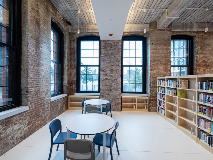 An interior shot of Adams Street Library, facing the windows. In front of the windows are two white tables with four blue chairs at each table.