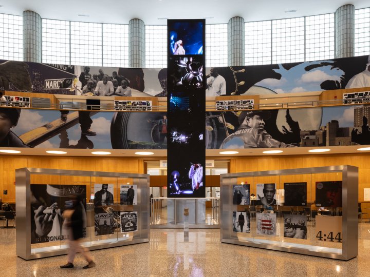 The Central Library lobby with various display cases showing JAY-Z photos and ephemera. In the center is a tower extending from floor to ceiling playing Jay-Z music videos. 