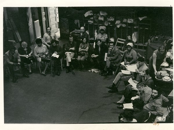 A group of 20 people sitting indoors on wooden chairs in an open circle. 