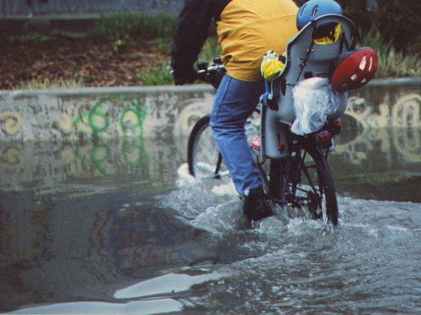 A person in jeans and a raincoat rides a bicycle on an urban street through 8 inches of water, a kid with a helmet sits on a rear seat. 