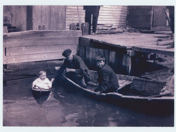 Black and white photograph depicting a toddler dressed in white in a small toy boat next to a skiff with a man and boy. The boat is next to a wooden dock and there is a person standing on the dock; only their legs are visible.