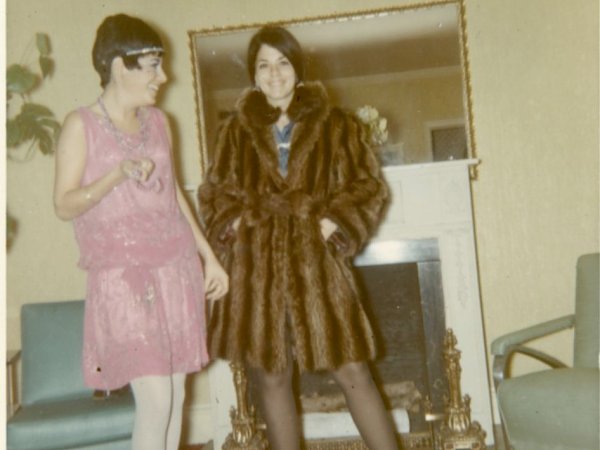 Two women stand in front of a fire place wearing 1960s fashion. The woman at the center of the photo is wearing a long, brown fur coat. The woman standing to her left is wearing a light pink, sparkly dress.