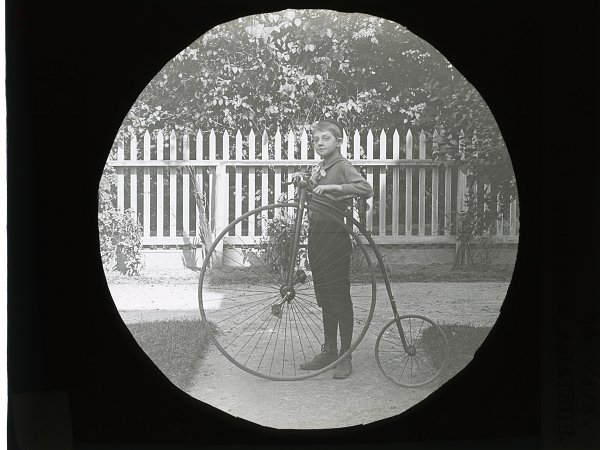 black and white photograph framed in a circle depicting a boy standing next to a penny farthing bicycle with a big wheel in the front and small wheel in the back. He is standing in front of a fence with trees and grass around. 